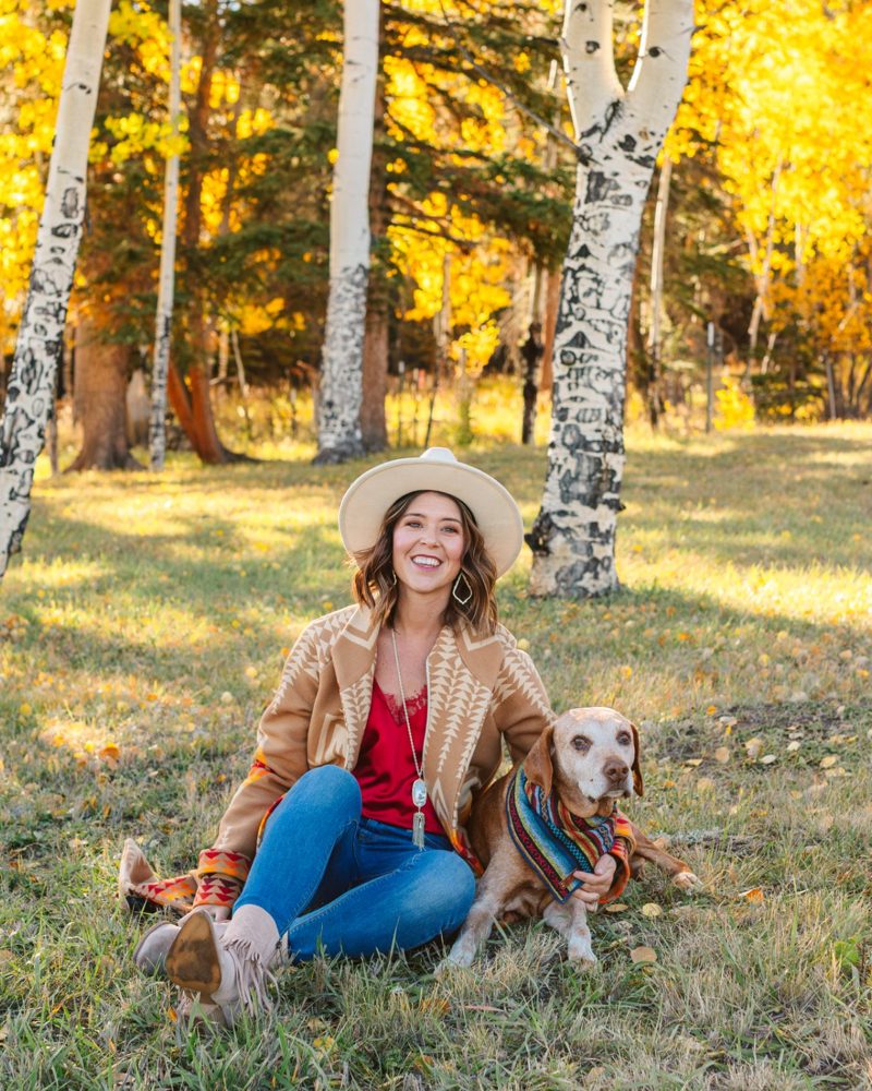 Danielle smiling in a cream hat and tan patterned coat, sitting in golden aspens with Ransom, her older dog with a white face, curled up beside her in a striped bandana