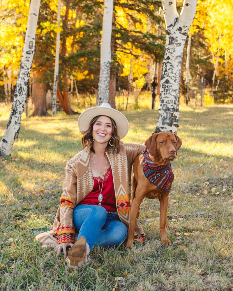 Danielle smiling in a cream hat and tan patterned coat, sitting in golden aspens with Reef standing beside her in a woven bandana